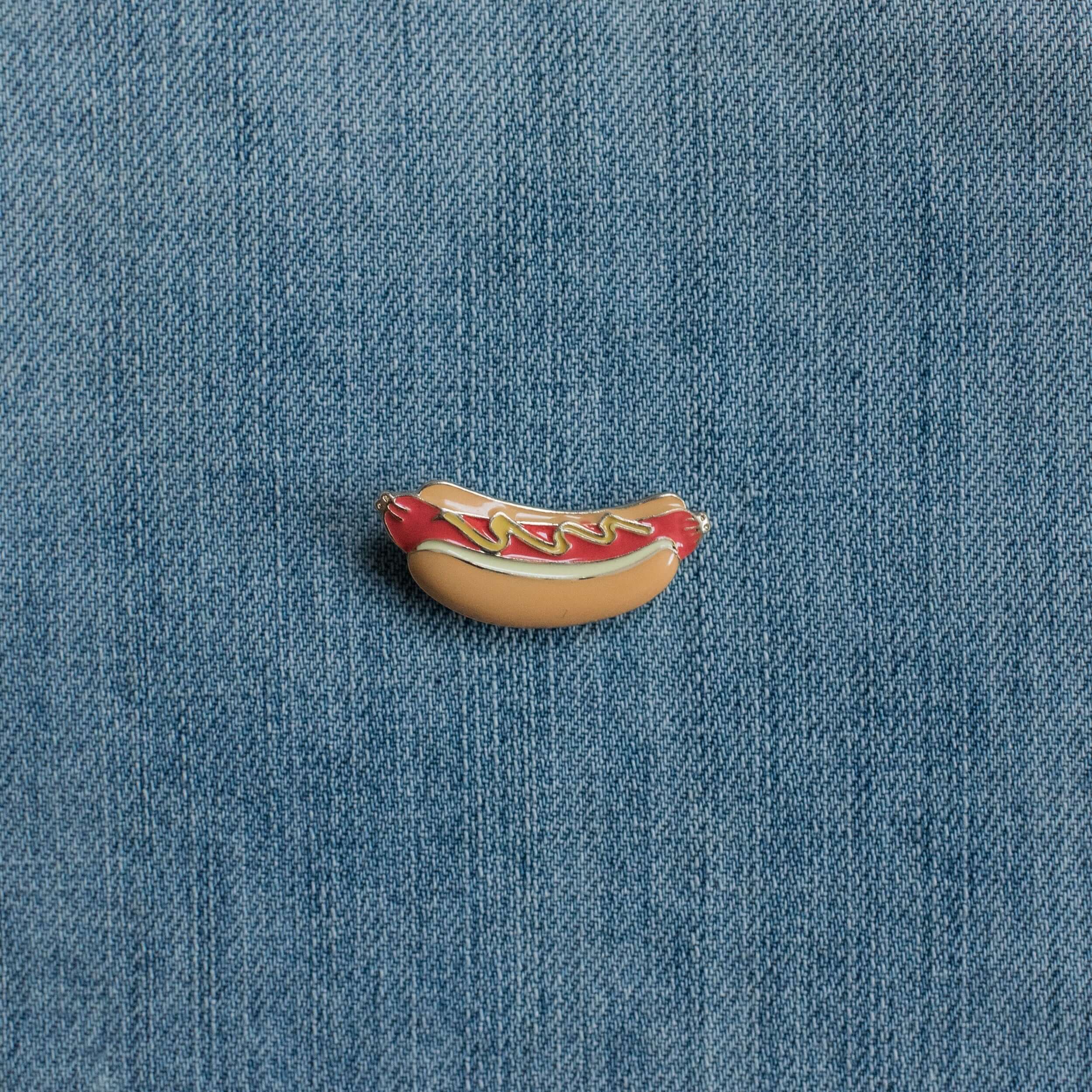 Hot dog enamel pin with ketchup and mustard on denim fabric background