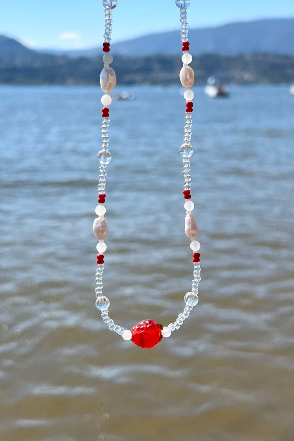 Handmade beaded necklace with red, clear, and white beads by the lake
