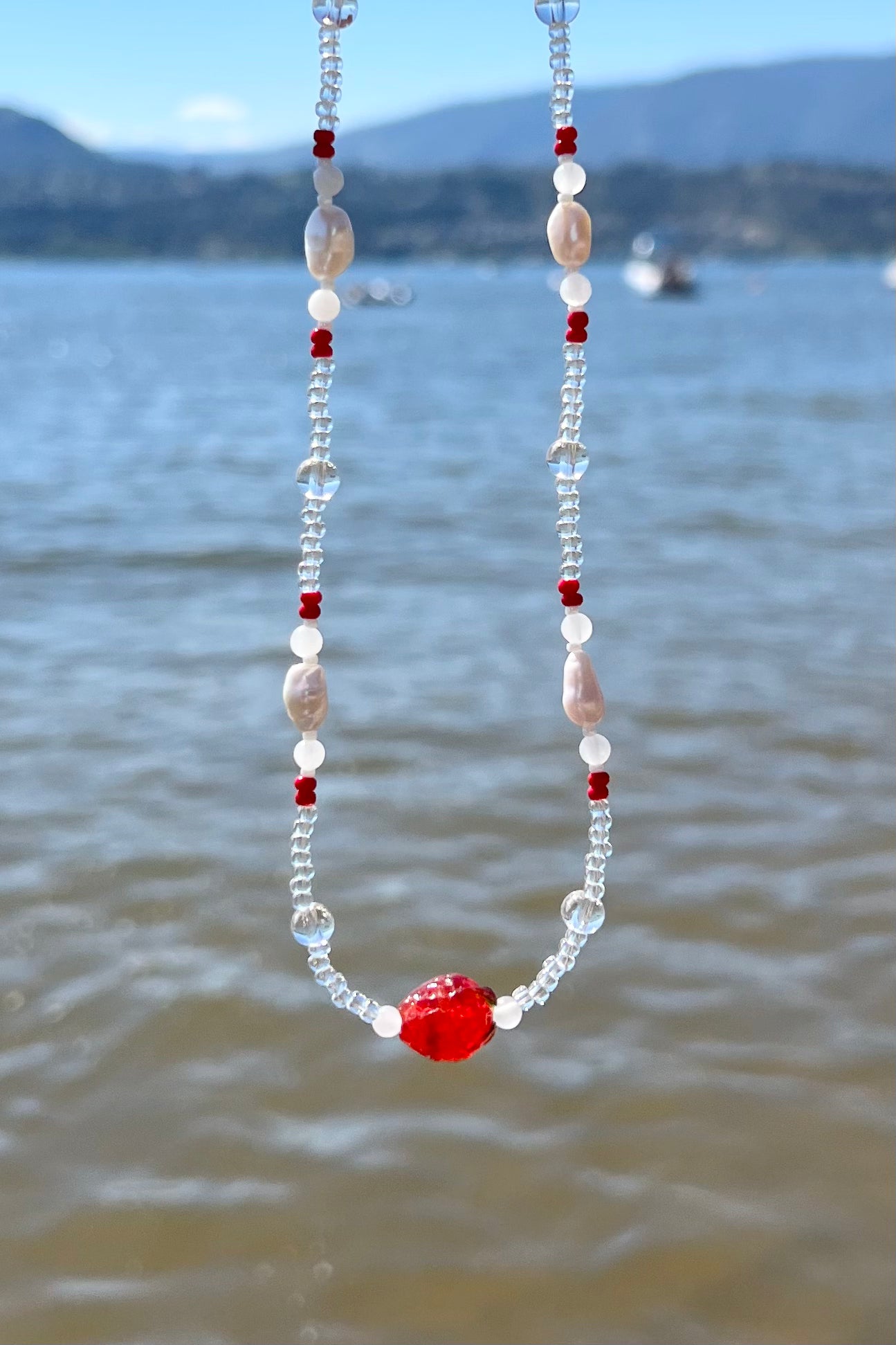 Handmade beaded necklace with red, clear, and white beads by the lake
