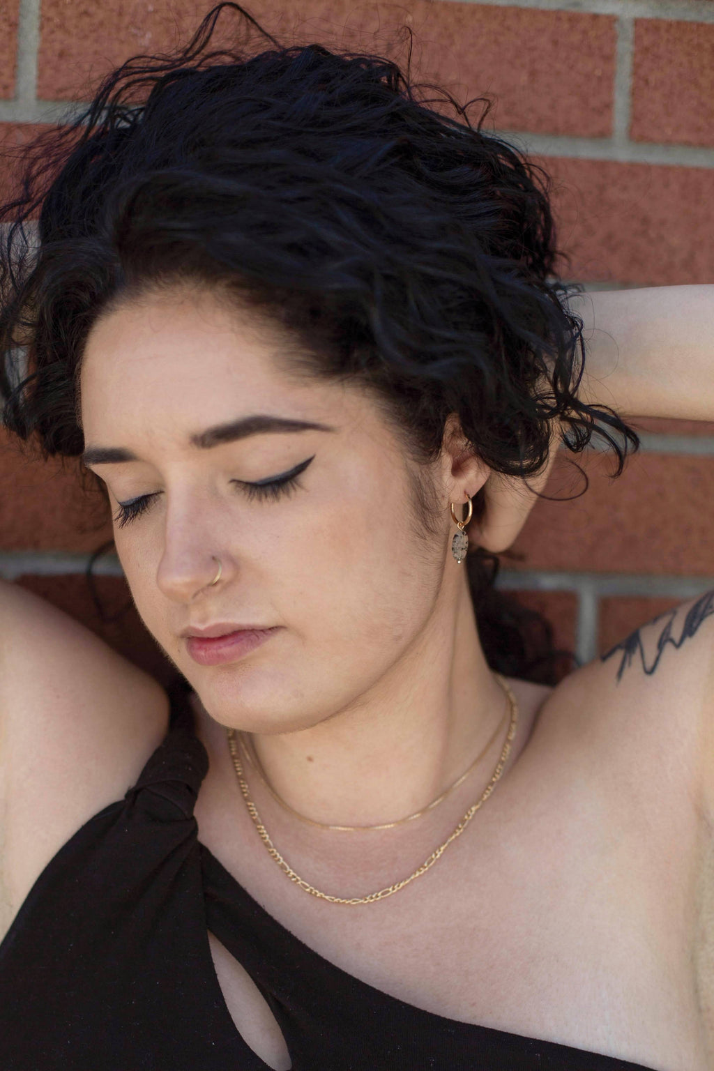Woman with curly dark hair and gold jewelry standing against a brick wall