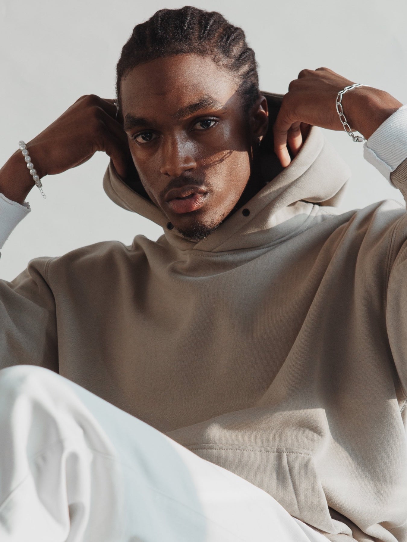Man in beige hoodie and white pants posing indoors, wearing silver and bead bracelets