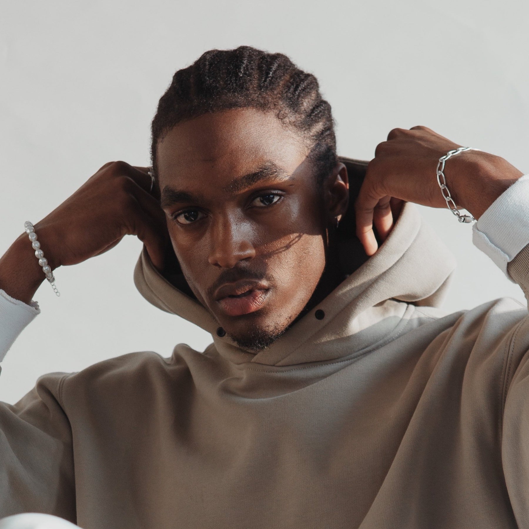 Man wearing beige hoodie, silver and pearl bracelets, neutral background, fashion portrait