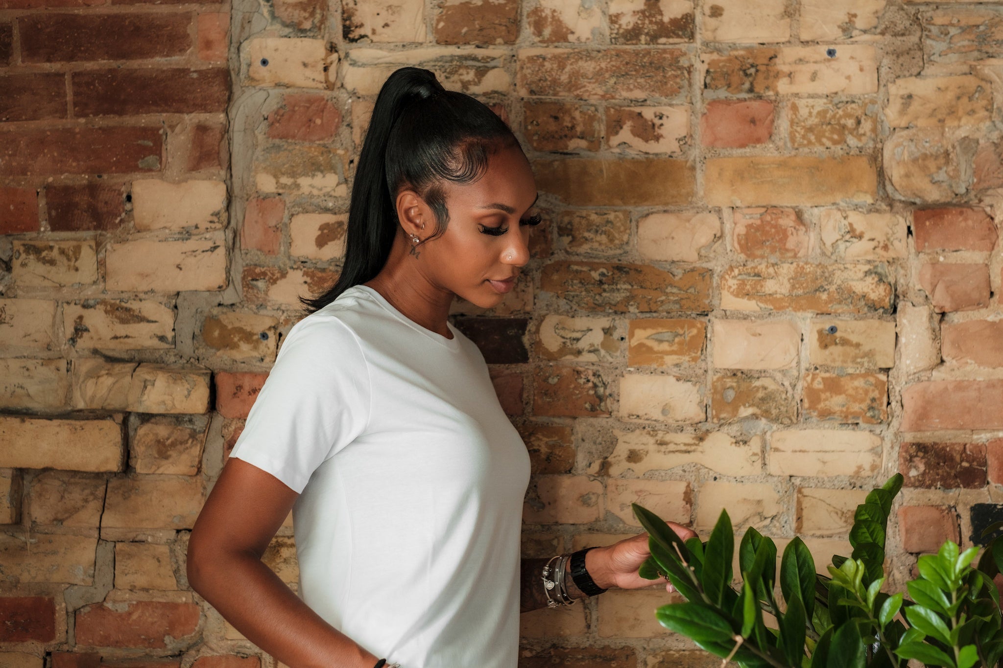 Woman in white shirt examining houseplants by rustic brick wall