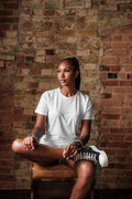 Woman in white t-shirt and sneakers sitting cross-legged in front of a red brick wall